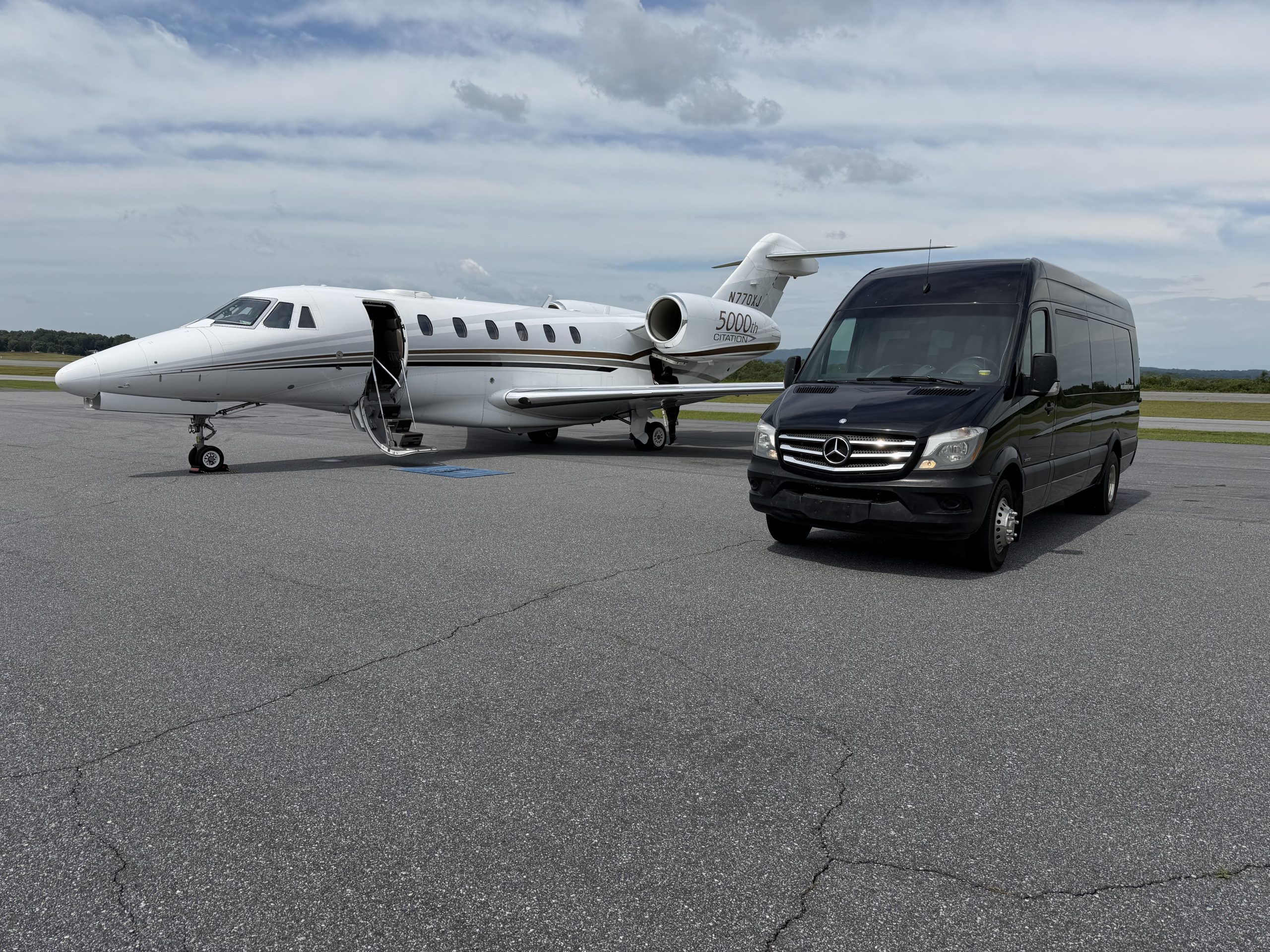 private-jet-sprinter-hickory-airport – Ashe County Livery blog White private jet with open door and portable stairs, parked on an airport tarmac beside a black Mercedes van under a cloudy sky.